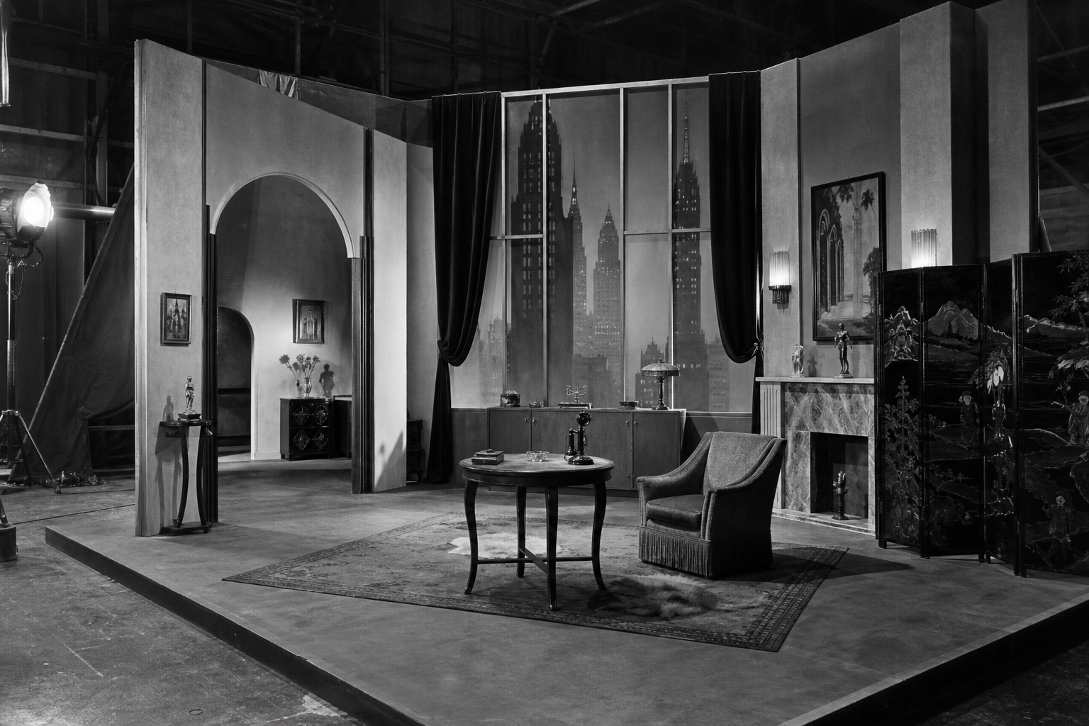 Black-and-white generated film-set interior with art deco walls, a city skyline backdrop, a fireplace, a table, a chair, curtains, and a visible studio light.